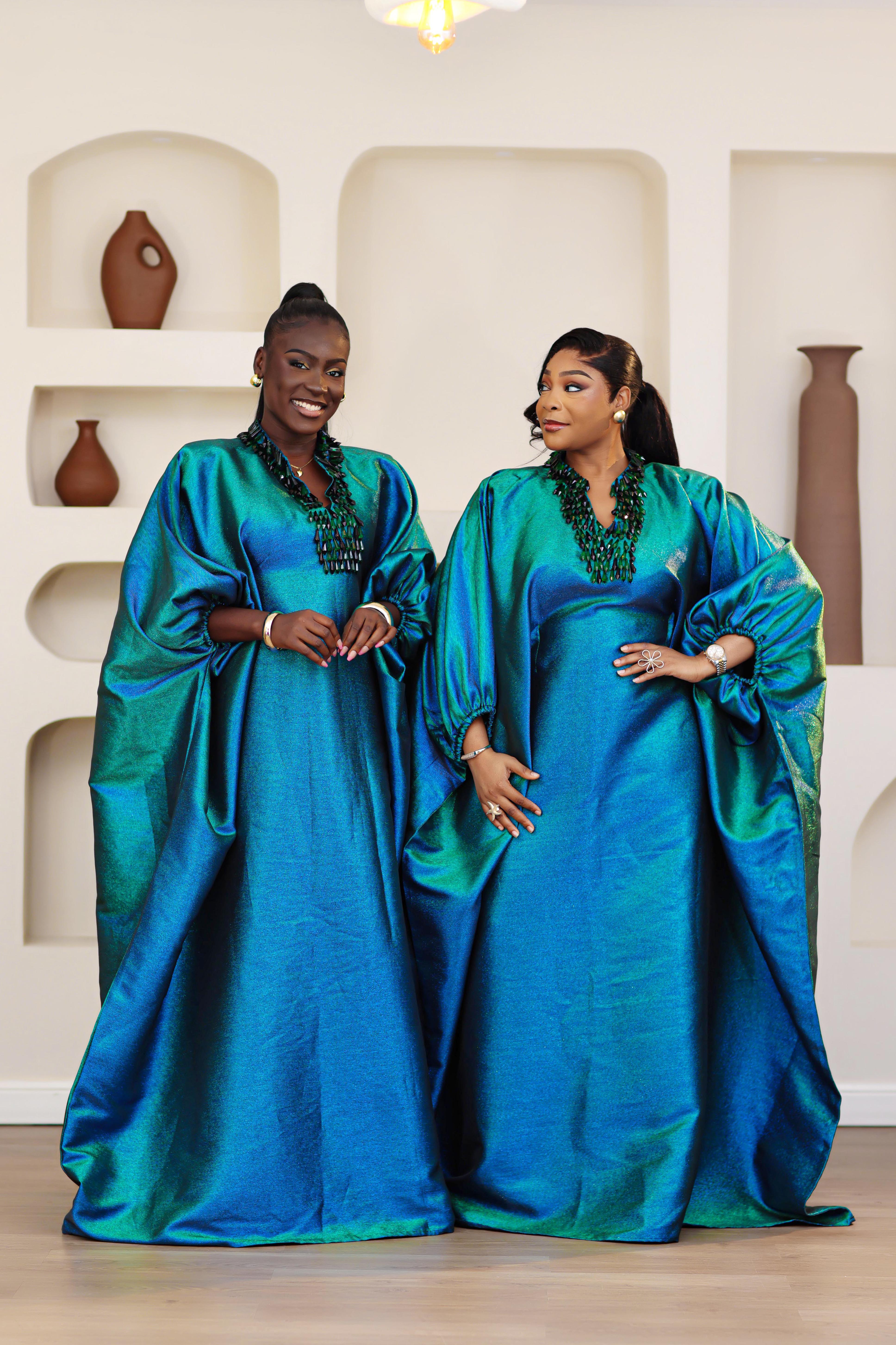 Women wearing a blue and green NIKI KAY Iridescent brocade boubou dress in a room with decorative vases on shelves.