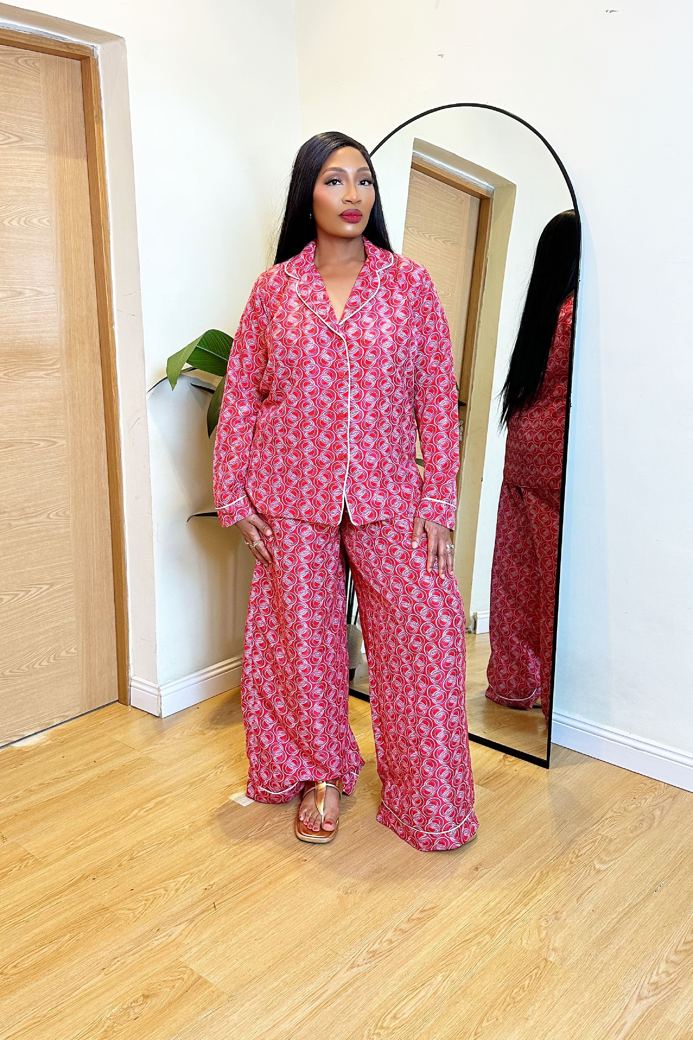 Woman in a colorful red pink African print Ankara pajamas shirt and pants set outfit standing in front of a mirror in a room with wooden flooring.