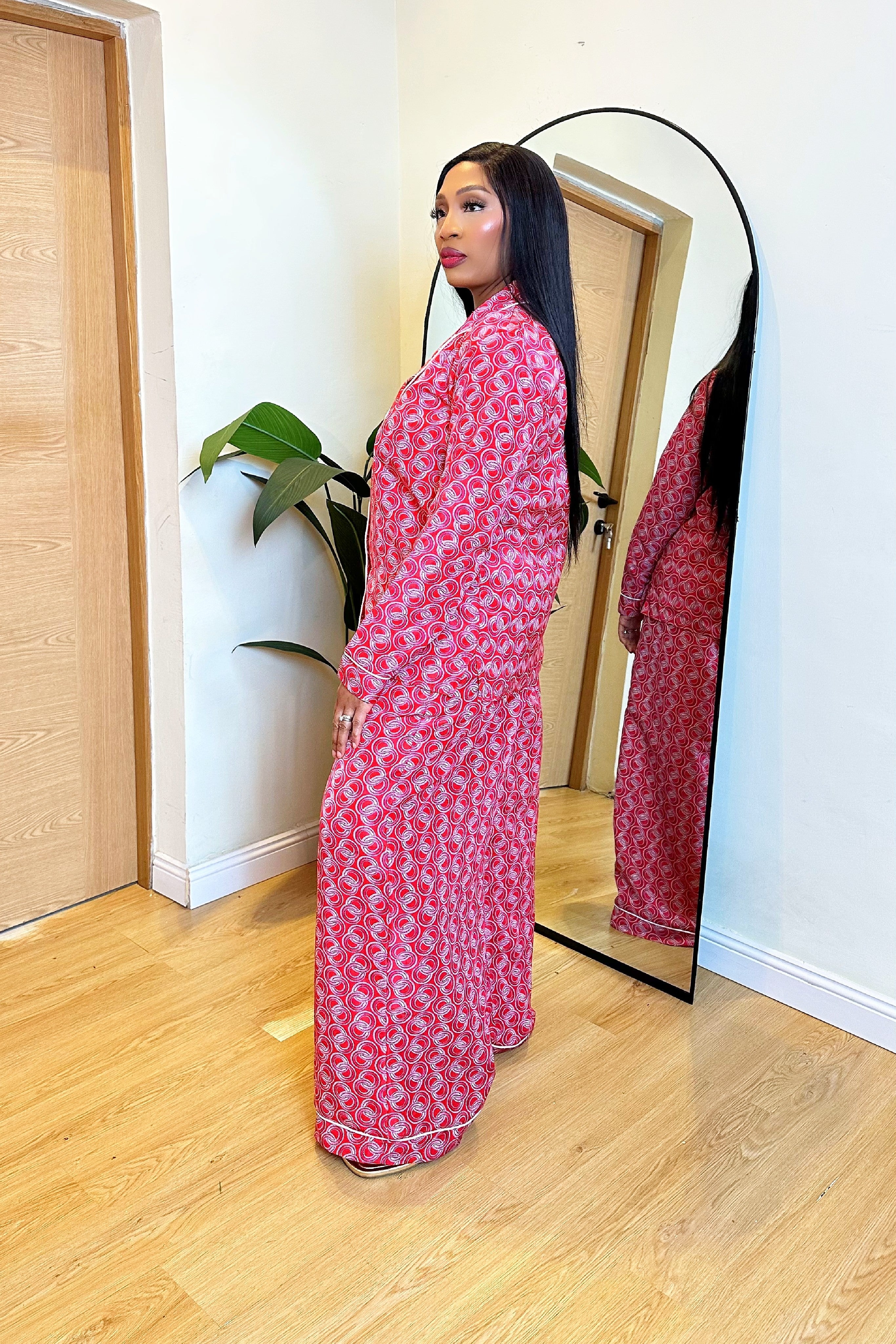 Woman in a colorful red pink African print pajamas shirt and pants set standing in front of a mirror in a room with wooden flooring.