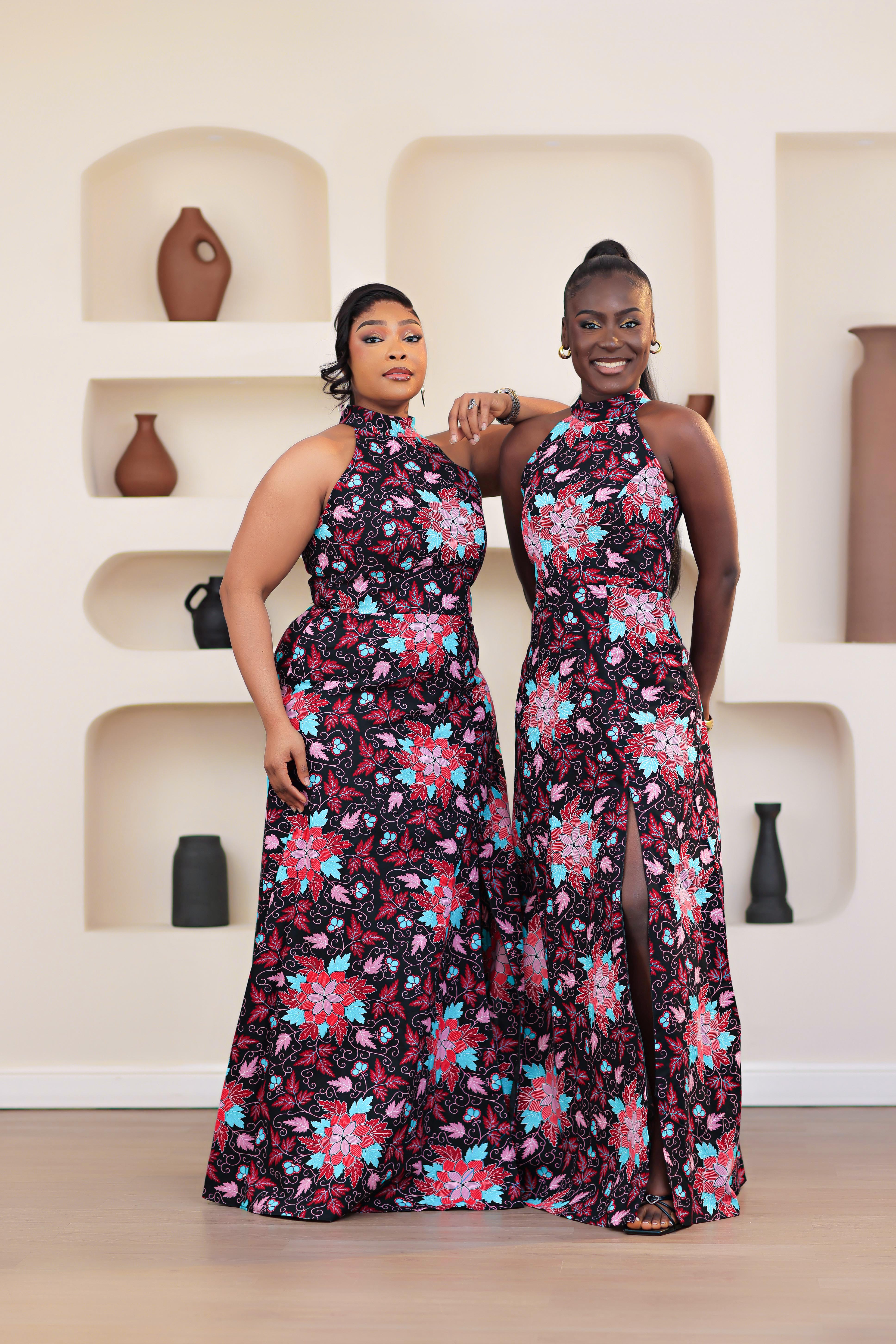 two women in an african print ankara pink multicolor floral dress posing in front of a decorative wall with shelves.