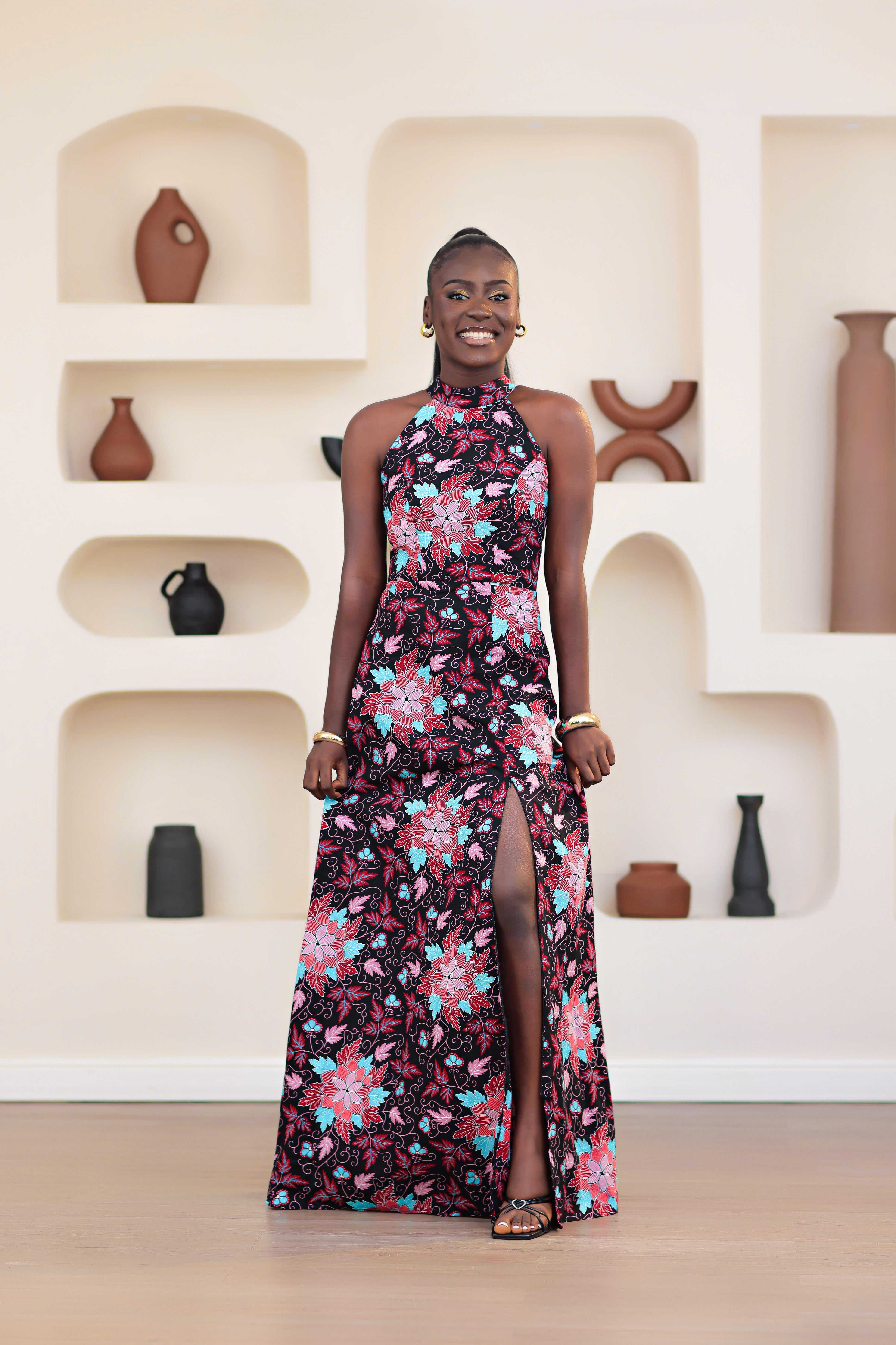 Woman in an african print ankara pink multicolor floral dress posing in front of a decorative wall with shelves.