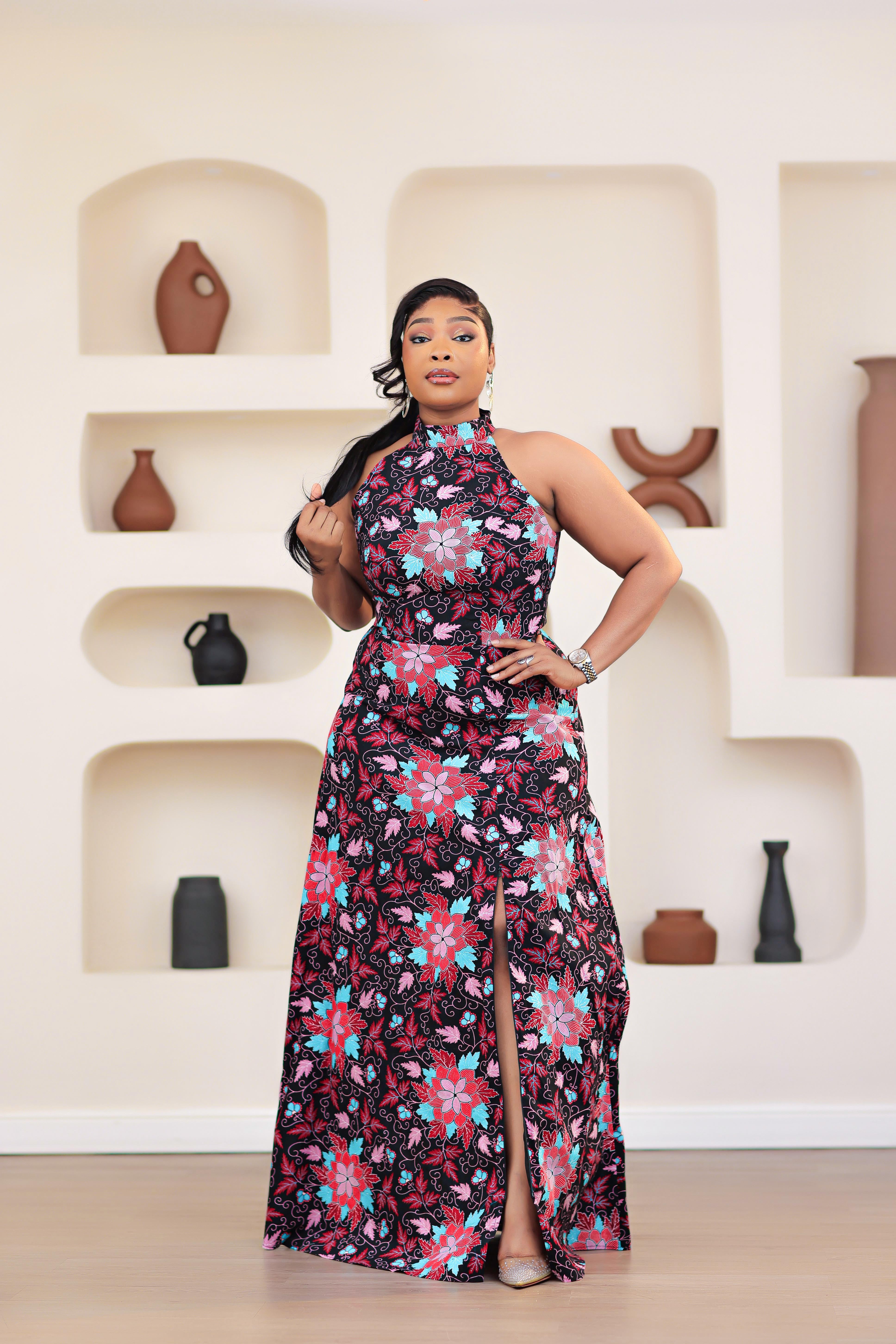 Woman in an african print ankara pink multicolor floral dress posing in front of a decorative wall with shelves.