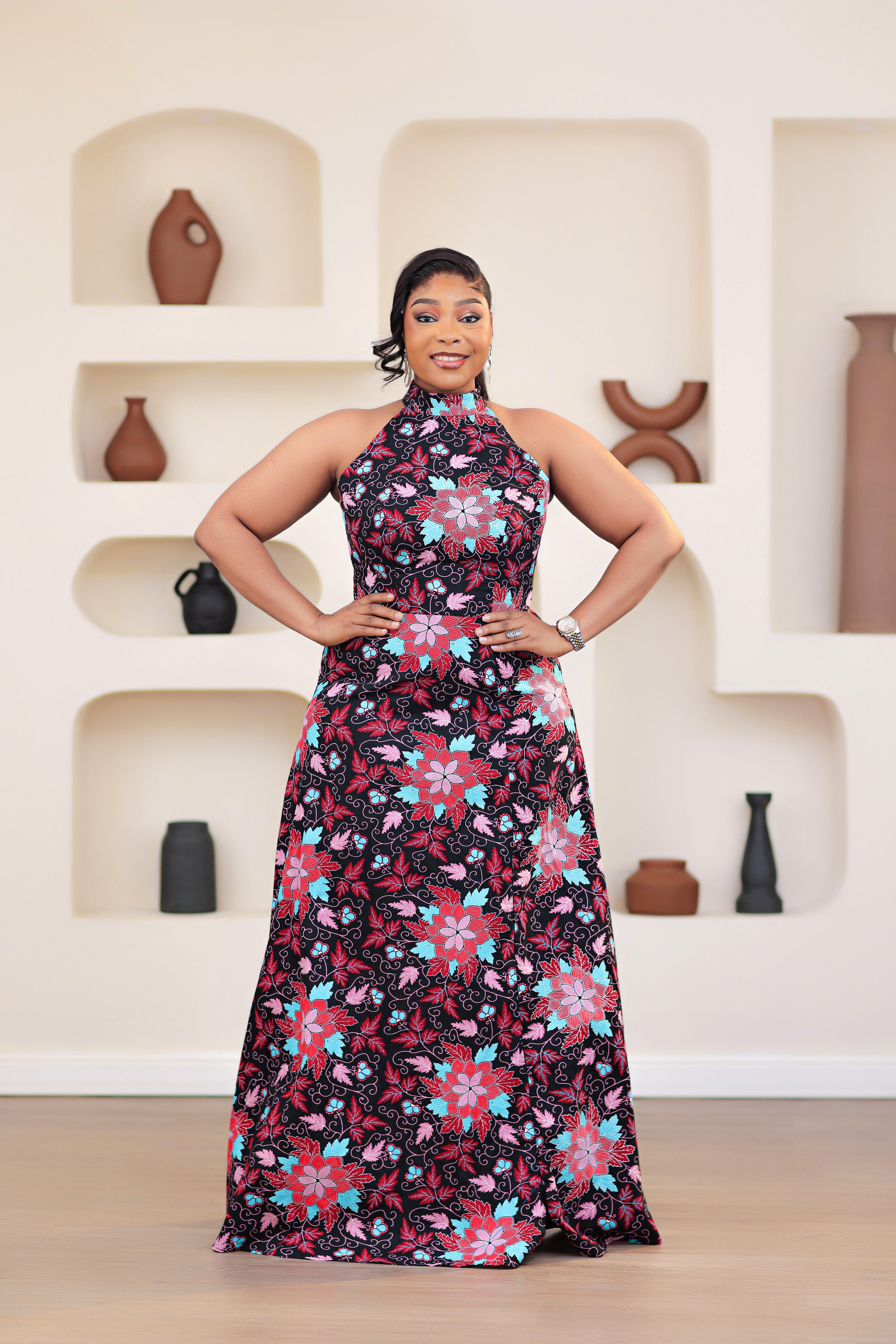 Woman in an african print ankara pink multicolor floral dress posing in front of a decorative wall with shelves.