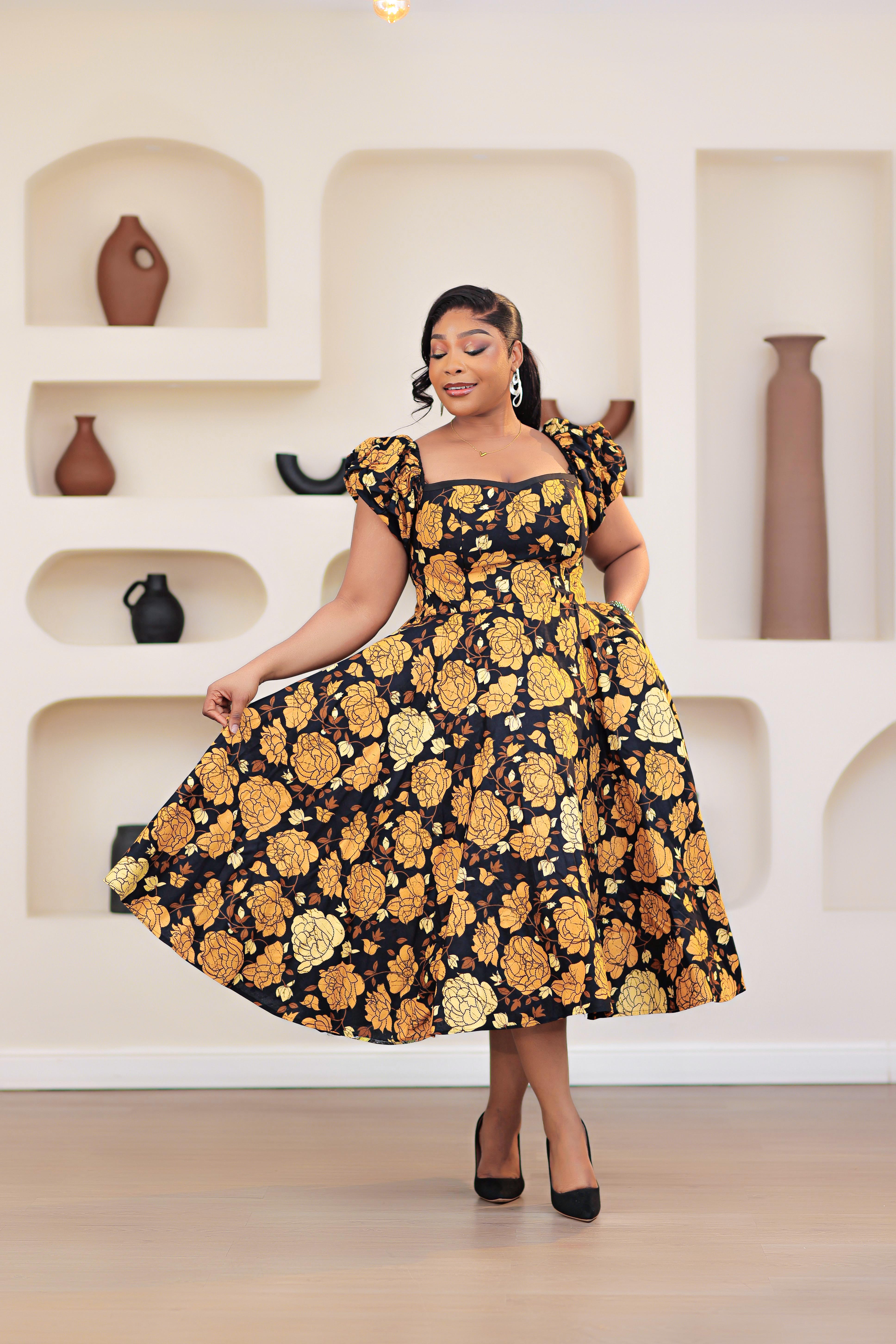Woman wearing an African print Ankara gold, brown, black, yellow short sleeve floral dress in a room with shelves and vases.