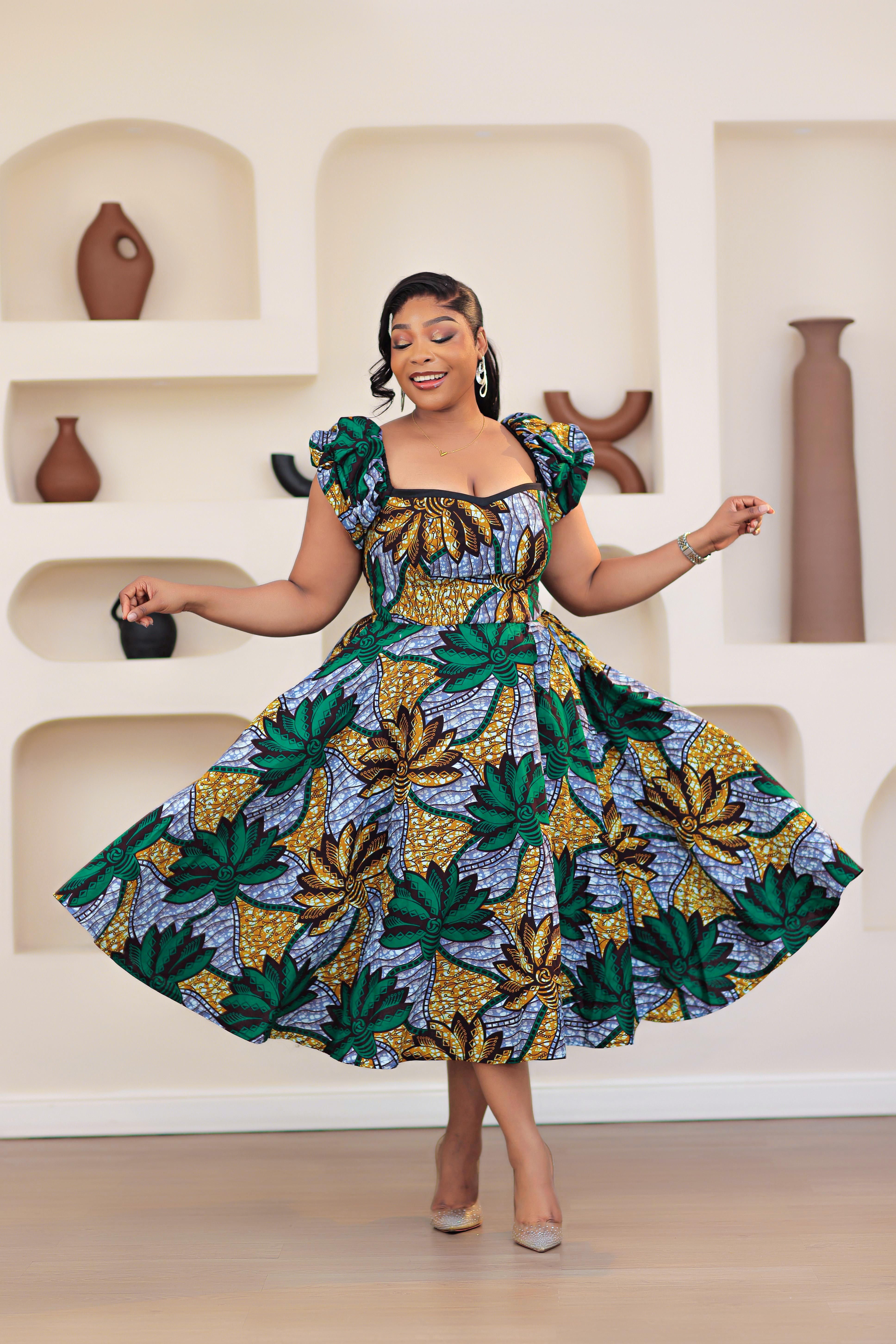 Woman wearing patterned African print Ankara green gold blue short sleeve floral dresses in a room with decorative shelves.