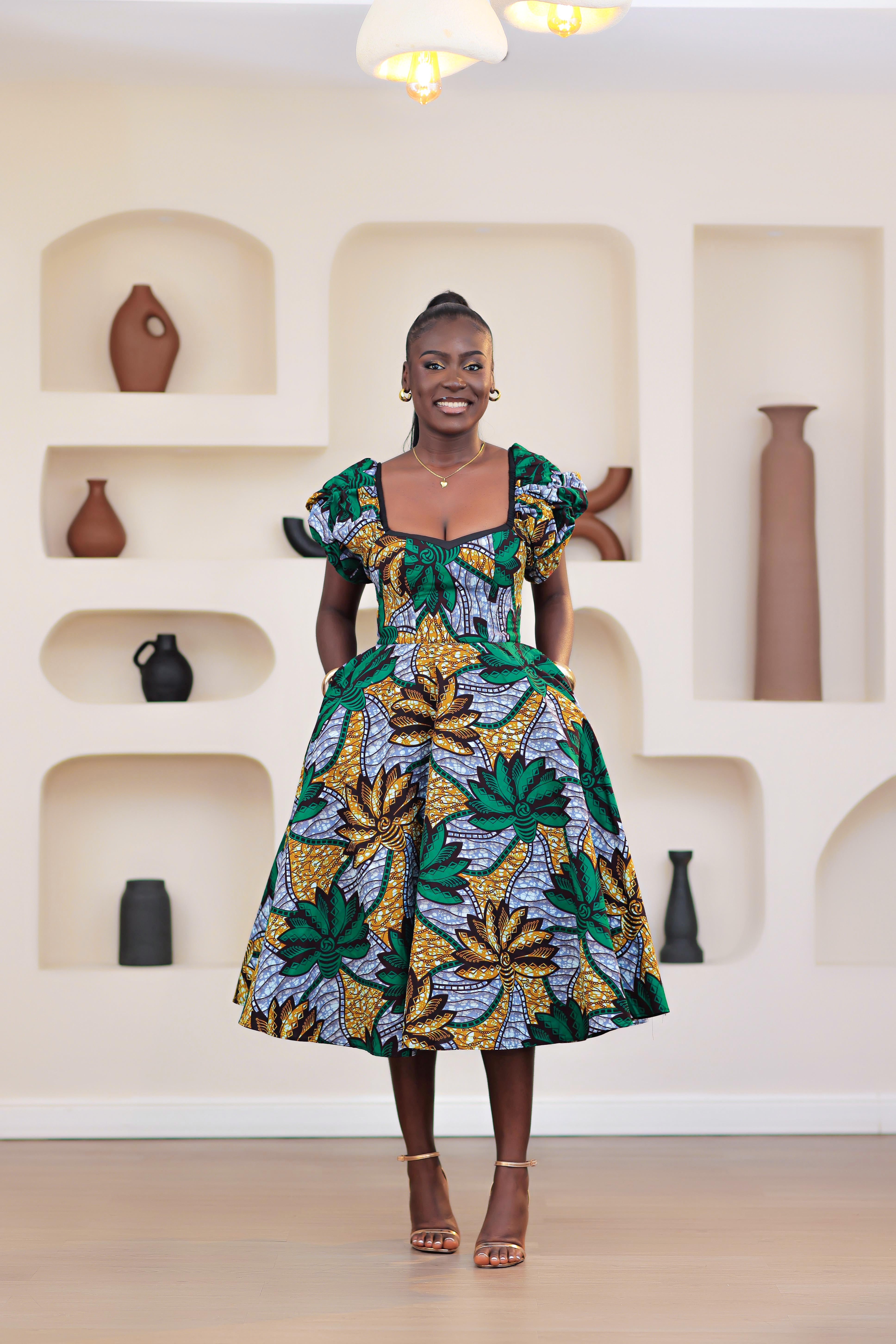 Woman wearing patterned African print Ankara green gold blue short sleeve floral dresses in a room with decorative shelves.