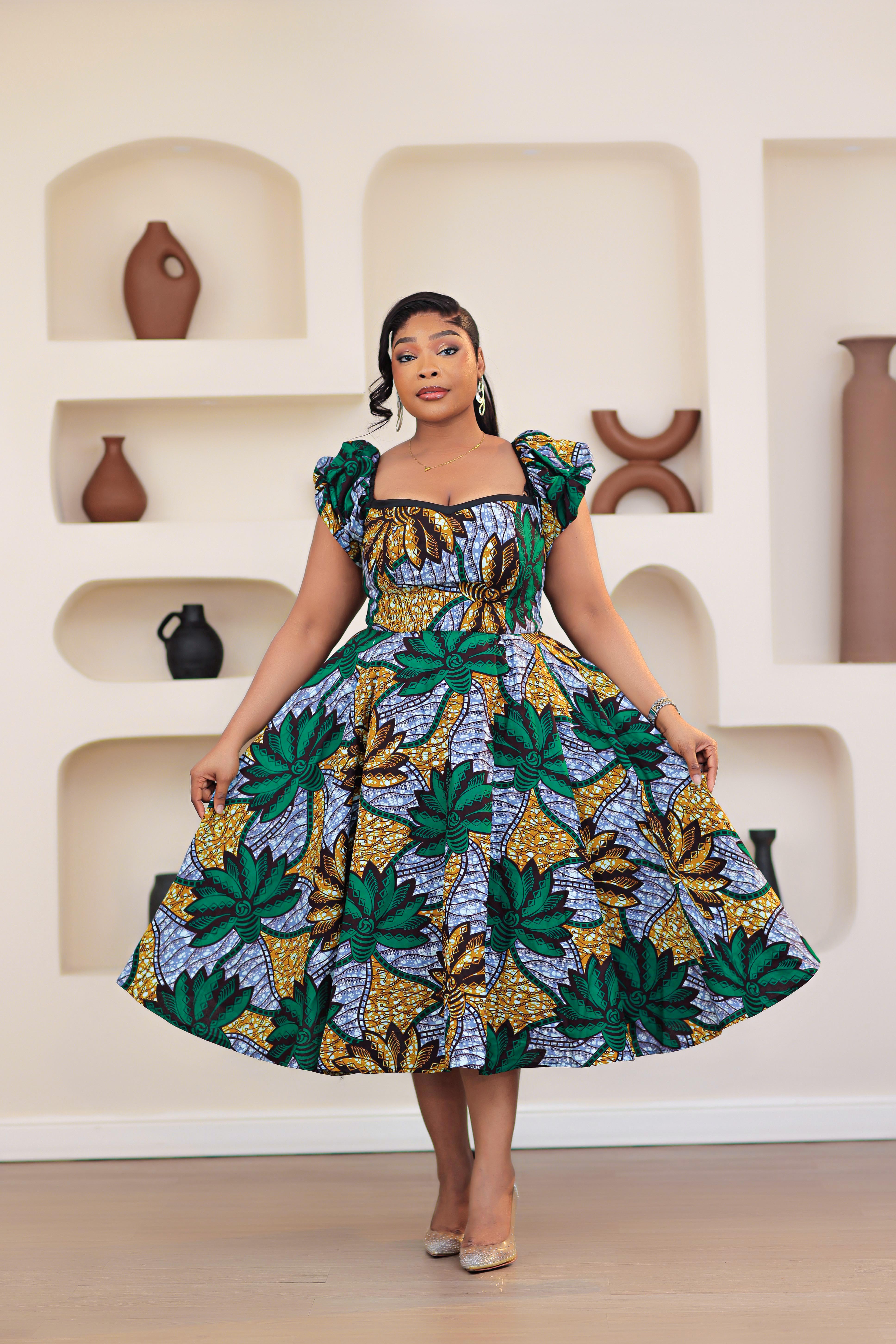 Woman wearing colorful patterned African print Ankara green gold blue short sleeve floral dresses in a room with decorative shelves.