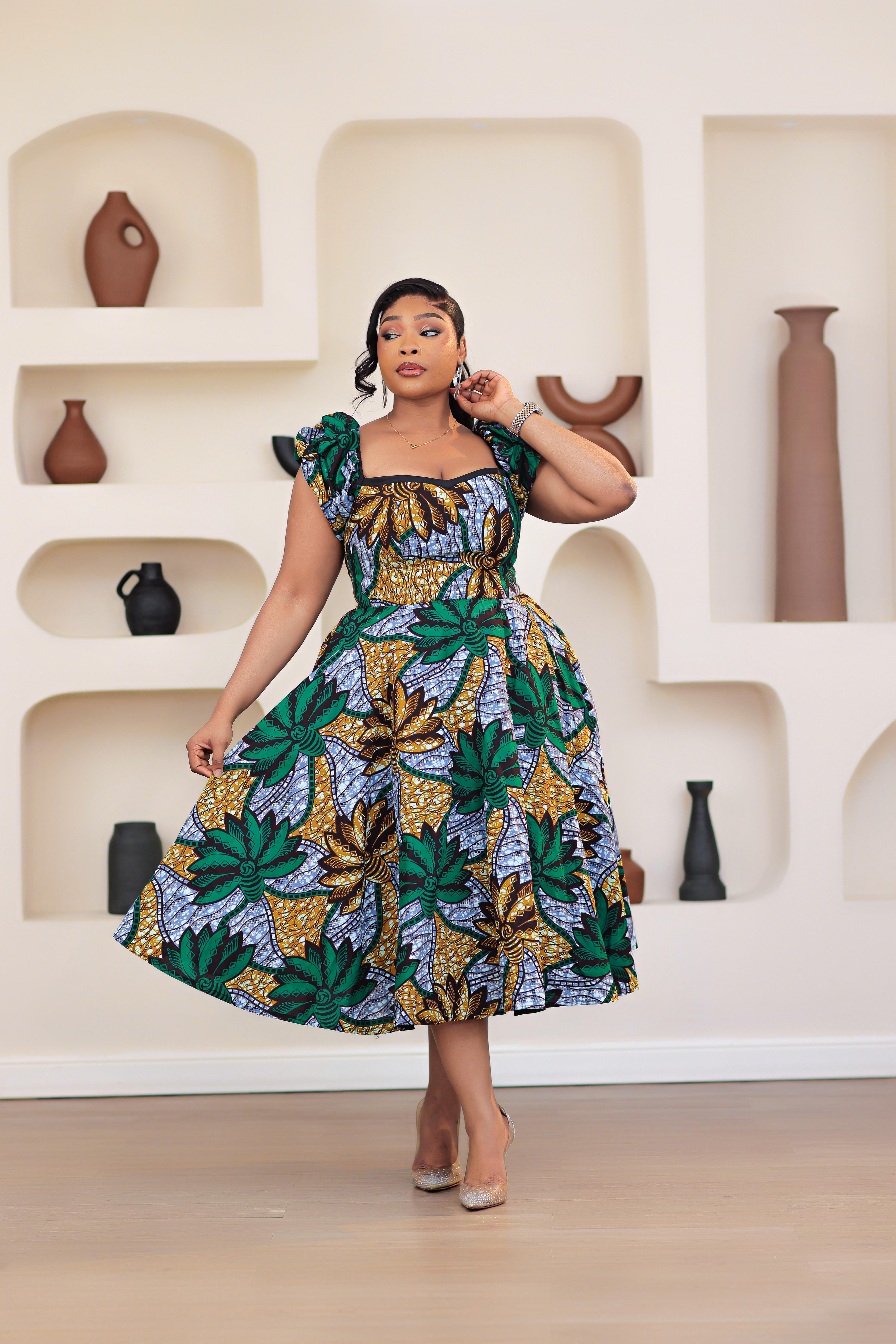 Woman wearing patterned African print Ankara green gold blue short sleeve floral dresses in a room with decorative shelves.