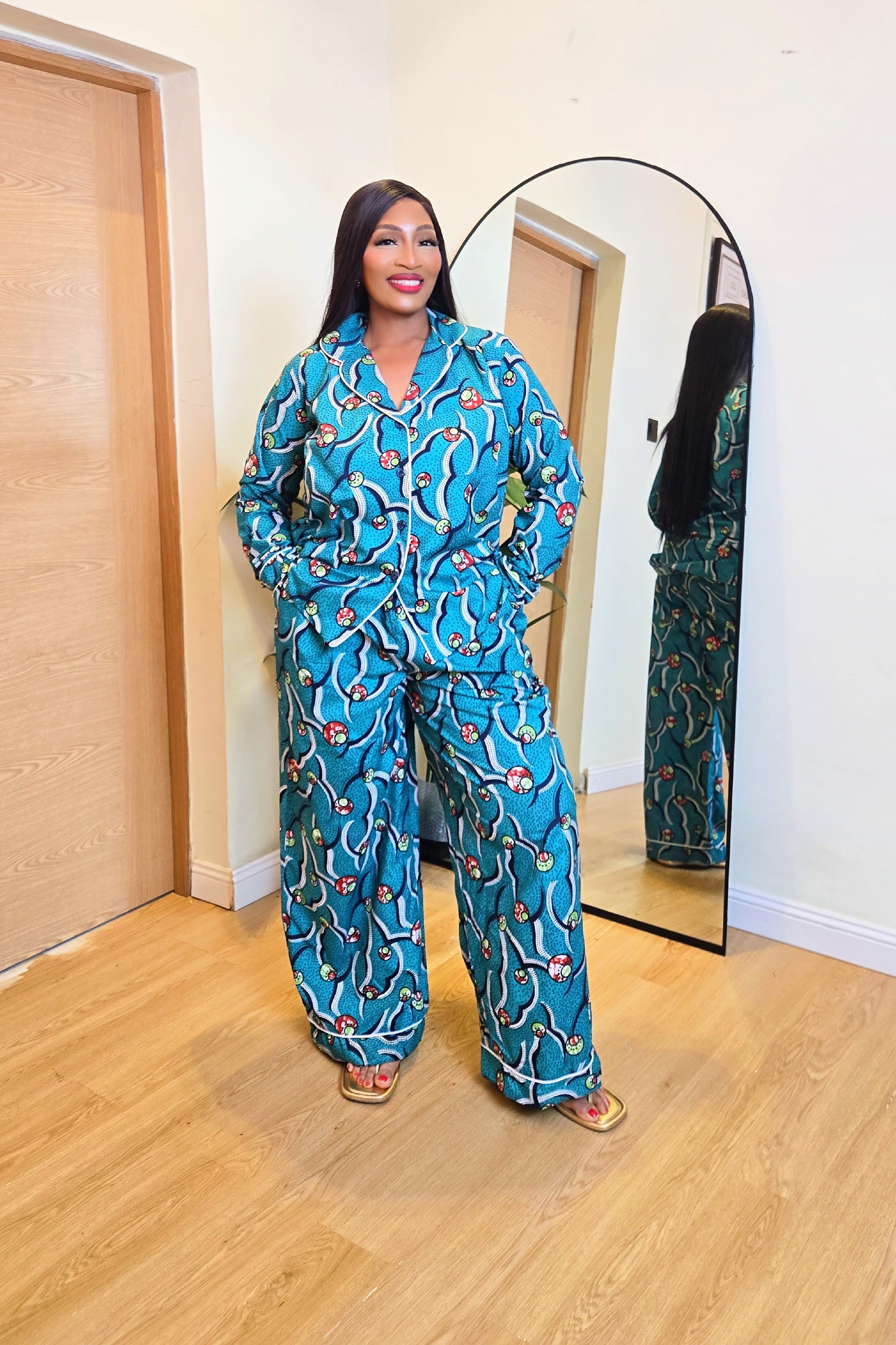 Woman in a colorful green teal African print pajamas shirt and pants set standing in front of a mirror in a room with wooden flooring.