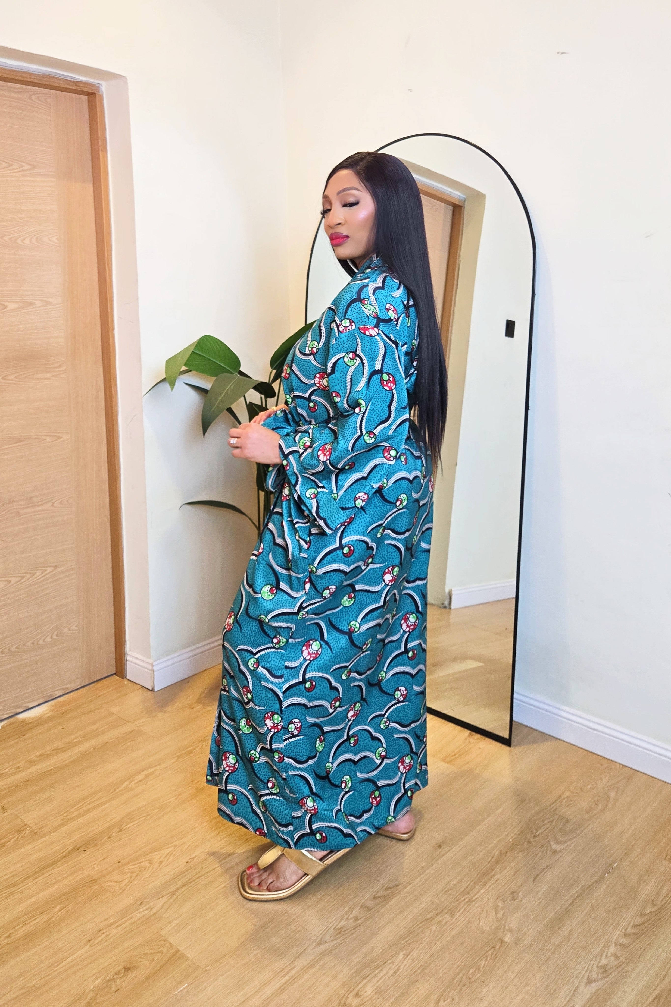 Woman in a colorful green teal African print robe dress standing in front of a mirror in a room with wooden flooring.