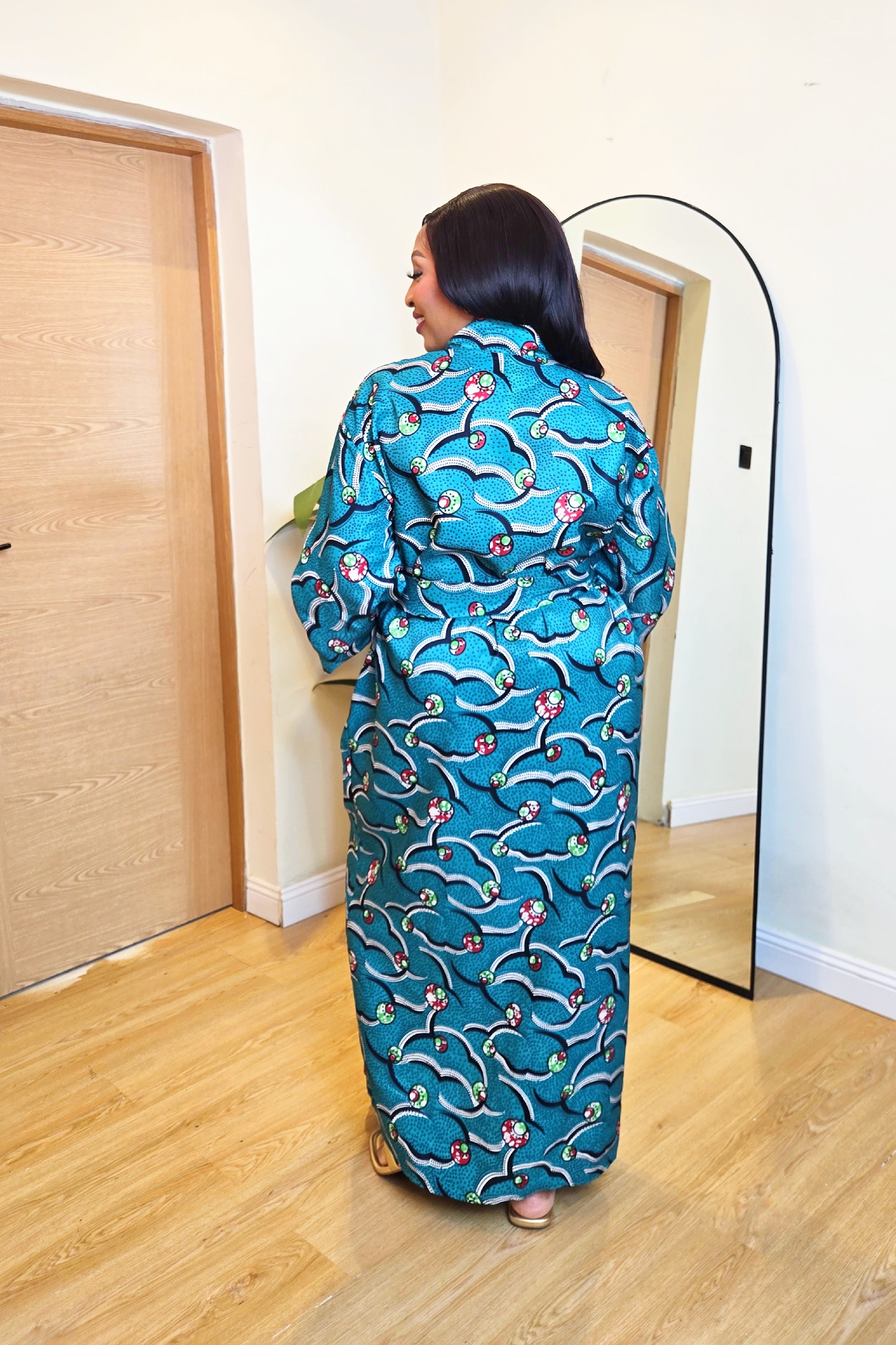 Woman in a colorful green teal African print robe dress standing in front of a mirror in a room with wooden flooring.